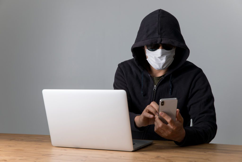 Mysterious person in a hooded jacket using phone and laptop.