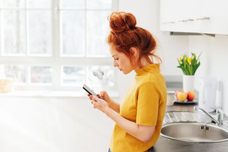 Woman in cute top hair bun using smartphone while leaning on kitchen sink