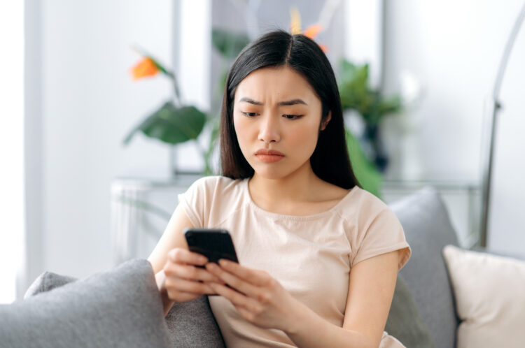 Upset Asian woman sitting on couch, using her smartphone