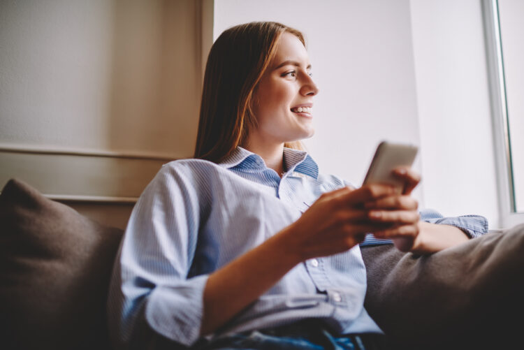 Thoughtful young woman resting after work at comfortable sofa with mobile phone in hand