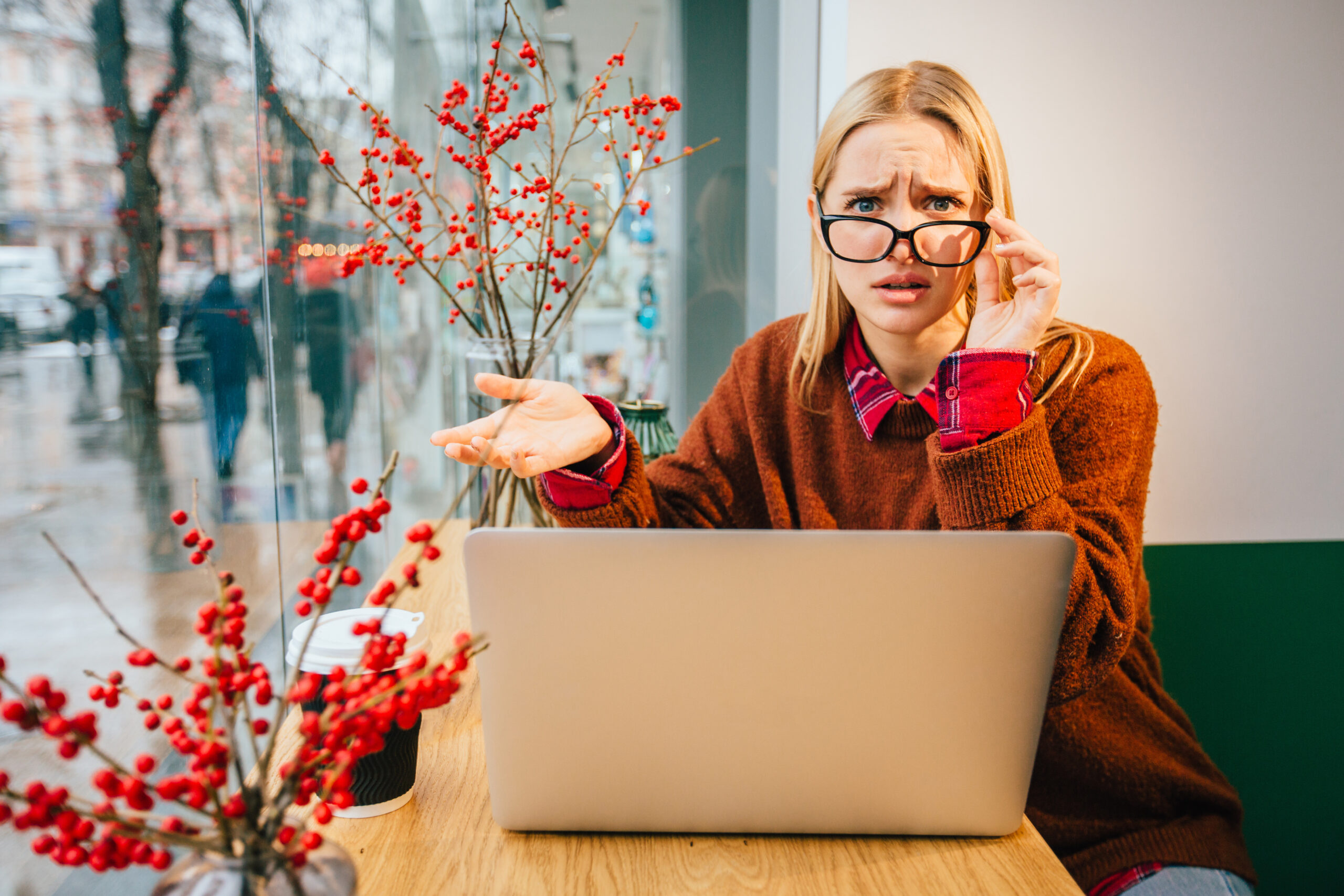 Young hipster business woman in eyeglasses with interrogative negative emotion wearing terracotta sweater, working with laptop near the window in modern interior cafe