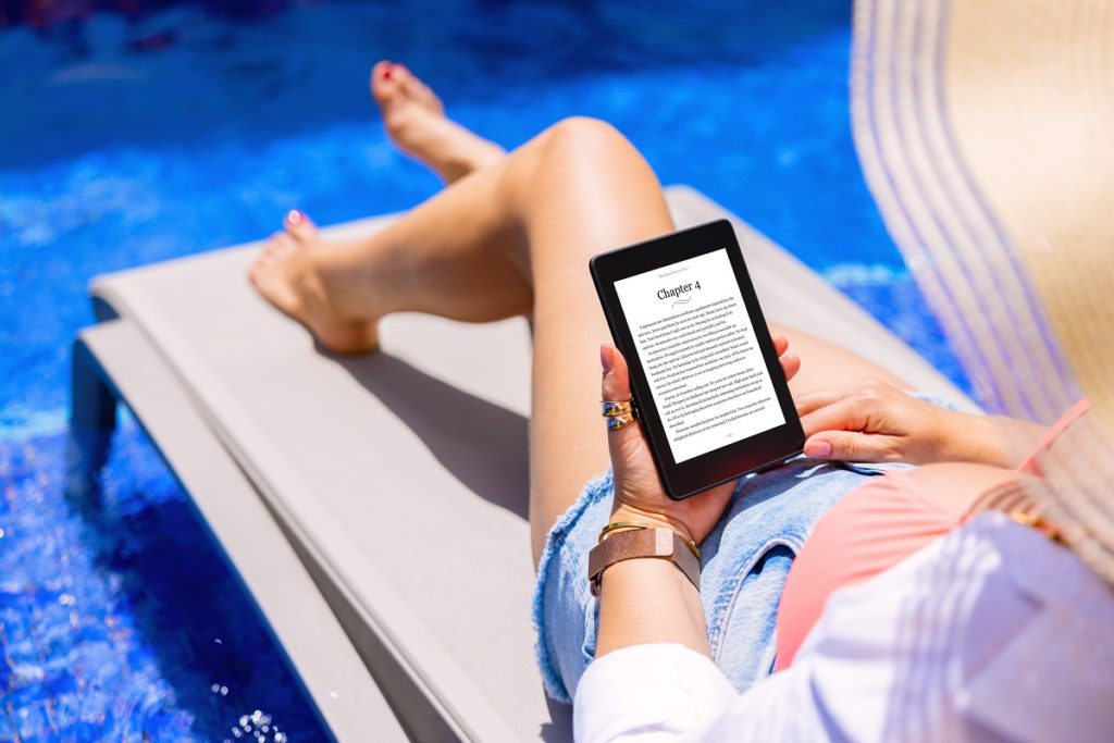 Woman holding e-reader device and reading e-book by the pool.