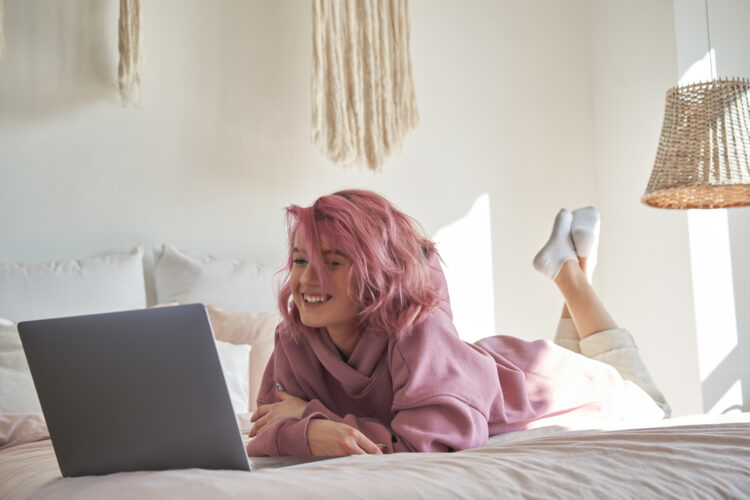 Happy teen girl with pink hair watching movie on laptop in bed.