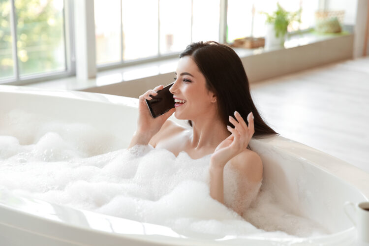 Relaxed young woman talking by mobile phone while taking bath in tub.