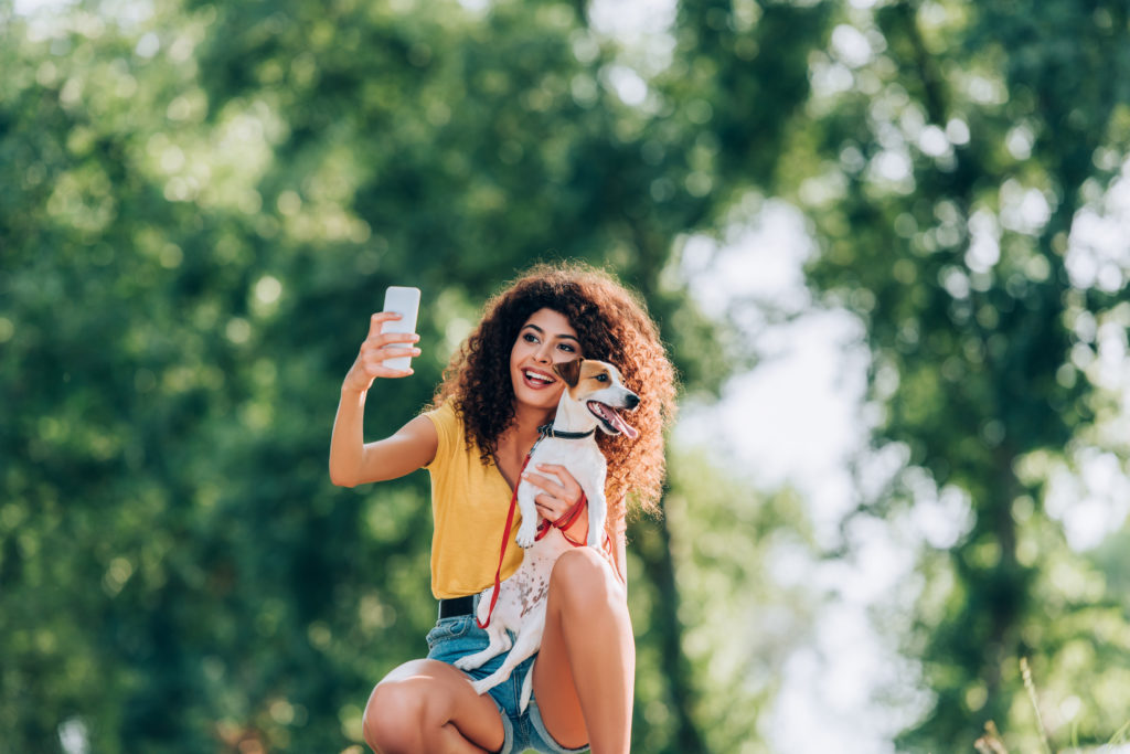 joyful woman in summer outfit taking selfie on smartphone with pet dog