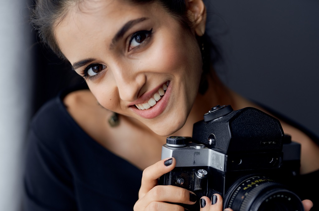 Brunette holding a camera near the window while posing elegantly for a picture.