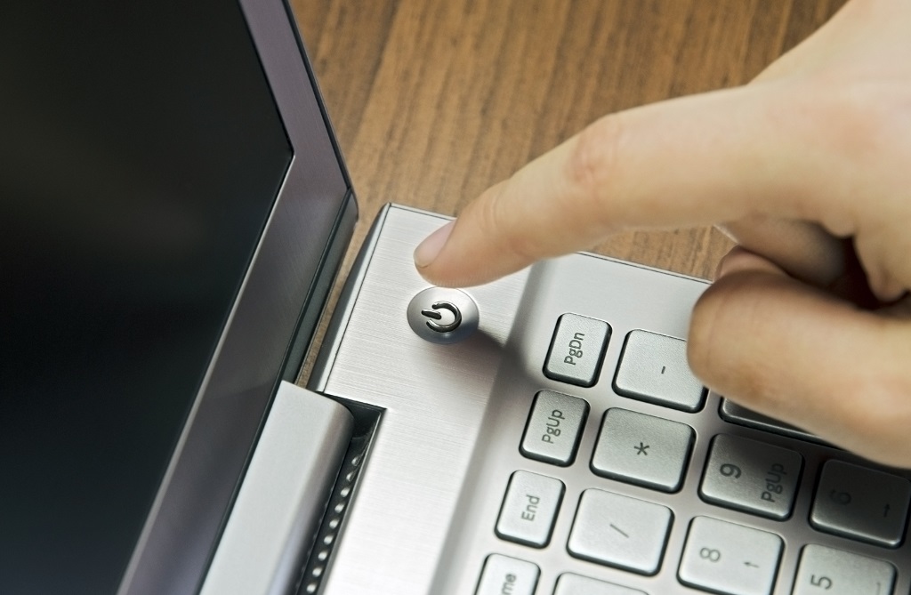 Female finger presses the power button on metallic silver laptop keyboard, lying on a wooden desk.