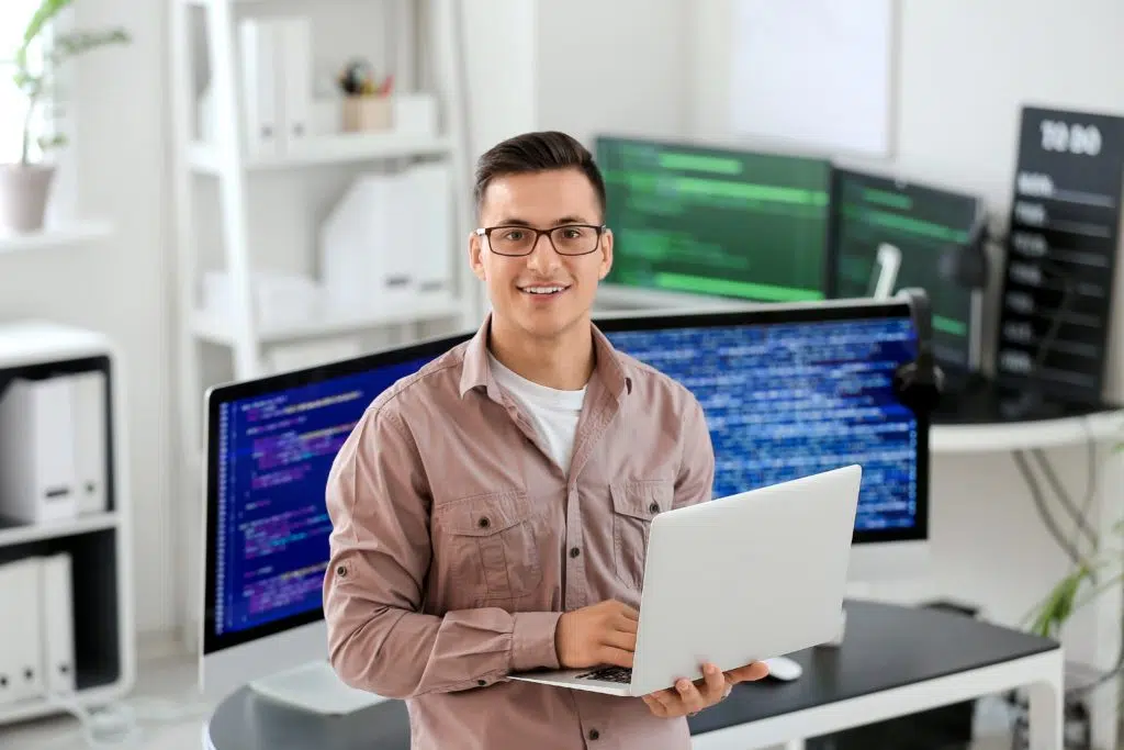 A programmer in glasses with laptop and computer monitors who understands the differences of IT vs ICT.