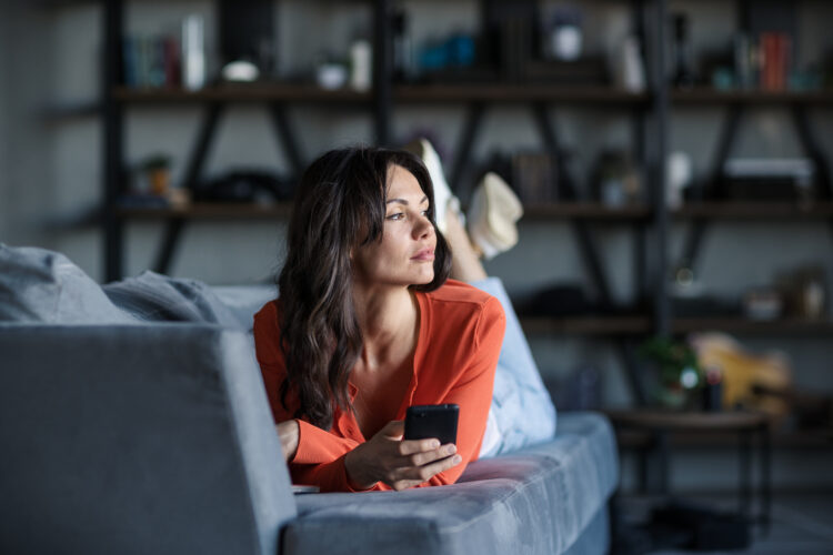 Woman thinking while texting on cell phone lying on sofa
