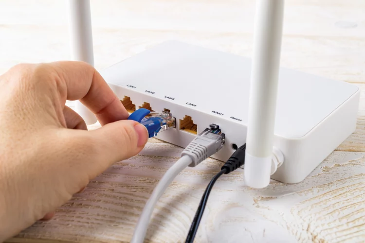 Woman hand plugging a blue network cable into socket of a white Wi-Fi wireless router on a white wooden table.
