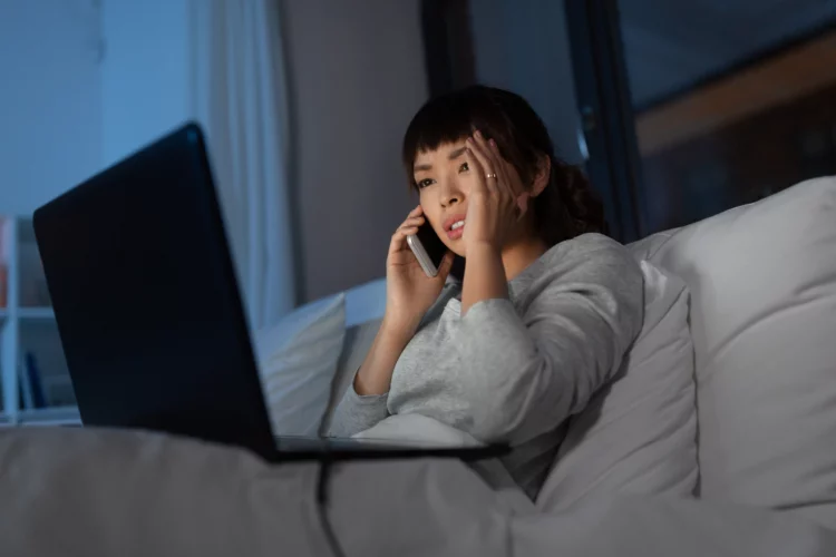 woman with laptop calling on phone in bed at night