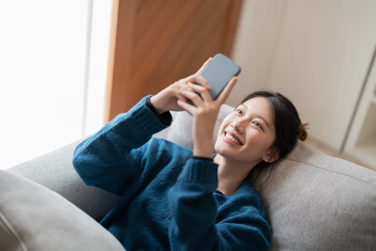 smiling attractive asian woman using mobile phone while lying on sofa at home