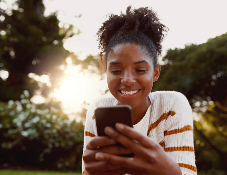 young afro woman using mobile phone in park on sunny day smiling 