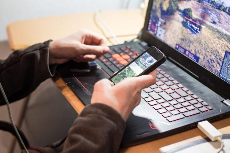 a young man holding a phone over laptop keyboard with video game on the screen
