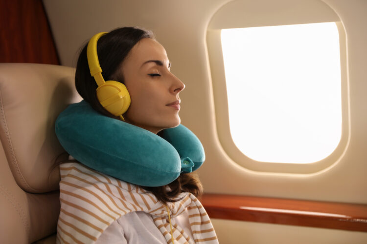 Young female traveler aboard aircraft resting head on travel pillow while listening to music