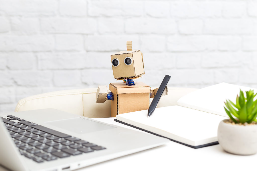 The robot sits at the desk and looks at the computer monitor.