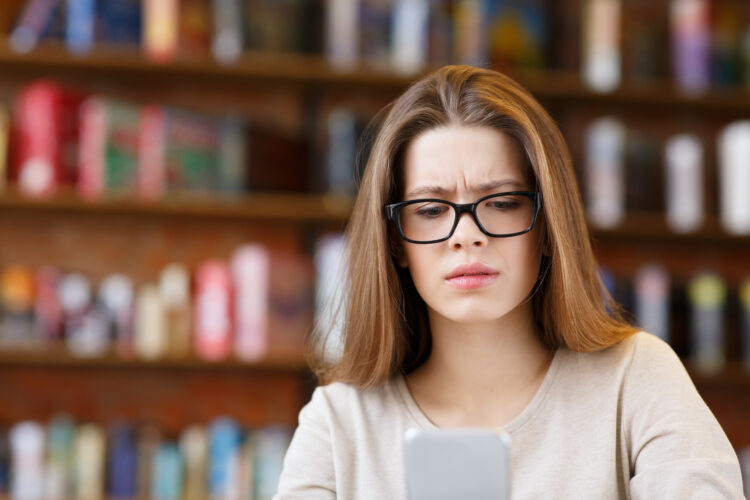 Young woman wearing eyeglasses sitting in cafe looking worried while looking at cellphone