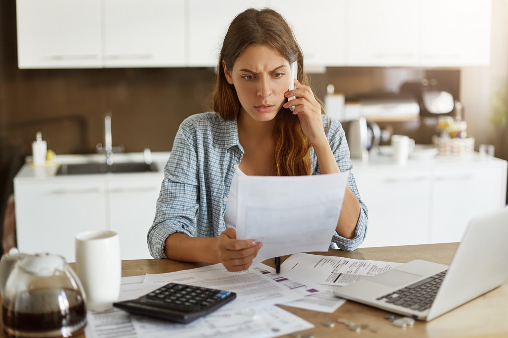 Worried woman dressed casually sitting at kitchen table, holding paper sheet while talking on the phone.