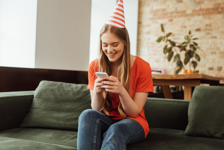 cheerful girl in party cap sitting on sofa and looking at smartphone