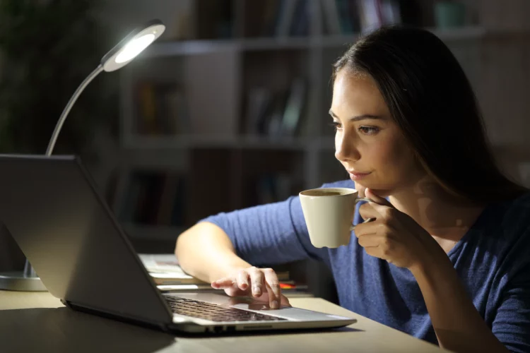 Woman using laptop holding coffee cup at night at home