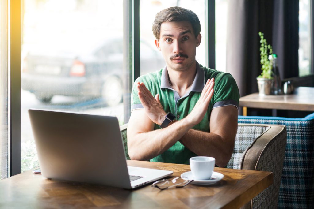 Serious man in front of his laptop, doing a "closed" sign with his arms.