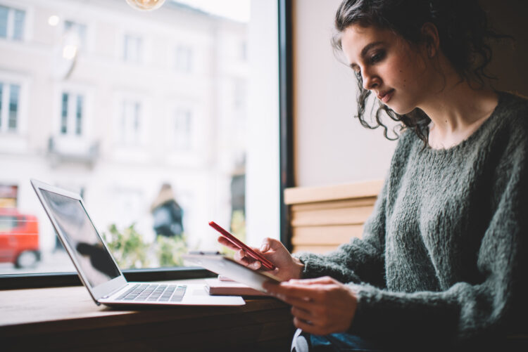 focused female freelancer browsing smartphone and reading notes in cafe