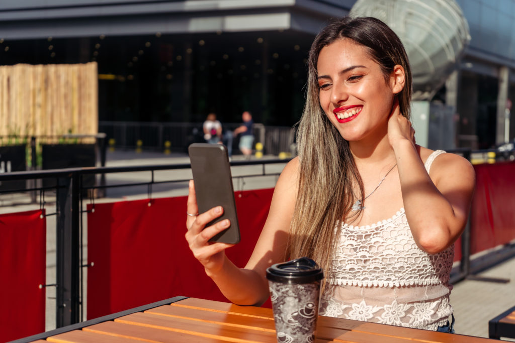 A beautiful woman sitting on the terrace of a bar having a drink while having a video chat on a sunny day.