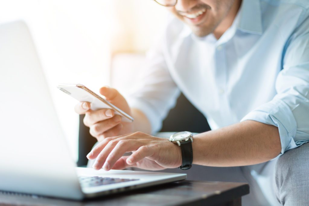 Smiling man typing on his laptop and cellphone.