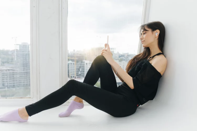 young woman with smartphone sitting on floor near window