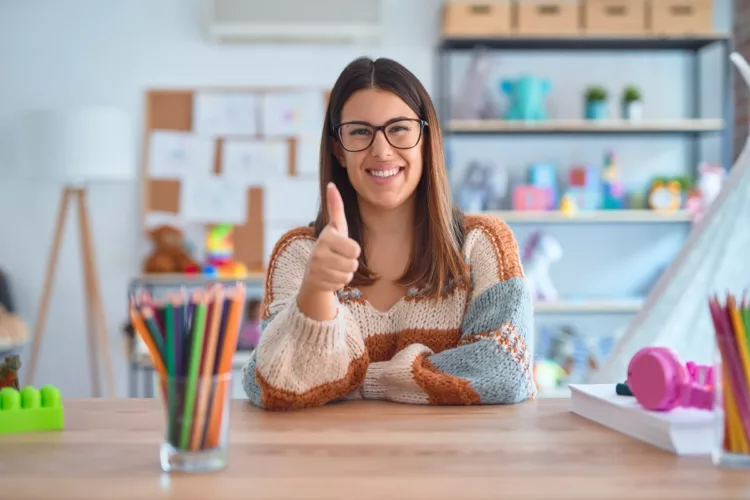 Young beautiful teacher woman wearing sweater and glasses sitting on desk doing happy thumbs up gesture