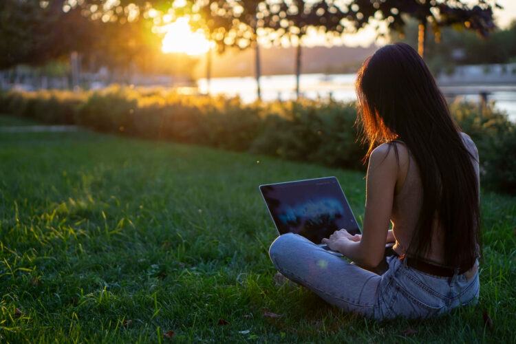 Young woman sits at sunset on the grass on school grounds and uses her laptop