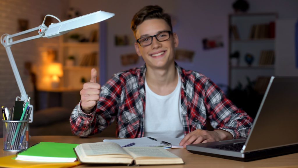 Student in glasses doing his homework while showing a thumbs-up sign to the camera.