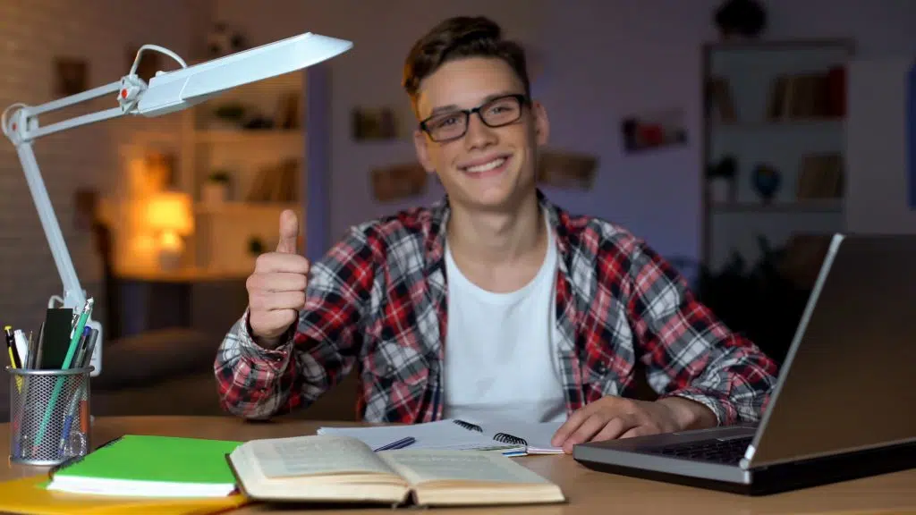 Student in glasses doing his homework while showing a thumbs-up sign to the camera.