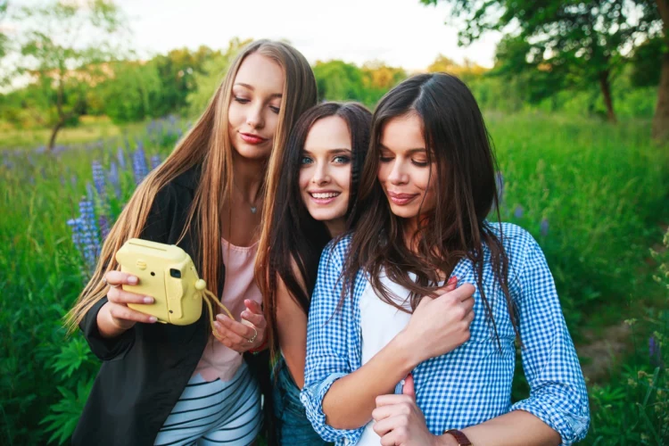 Three hipster girls blonde and brunette taking self portrait on polaroid camera and smiling outdoor. 
