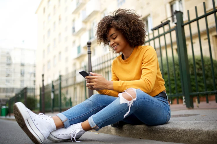 Young african american woman sitting outside on street using smartphone.