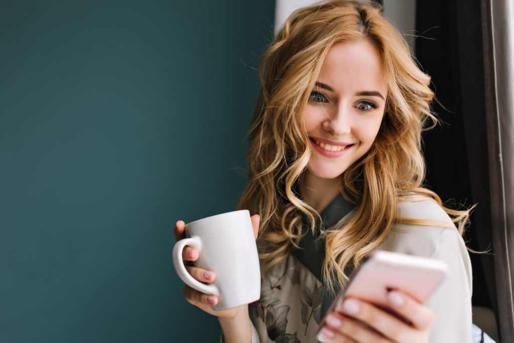 Beautiful young woman with cup on one hand her smartphone in the other looking excited.