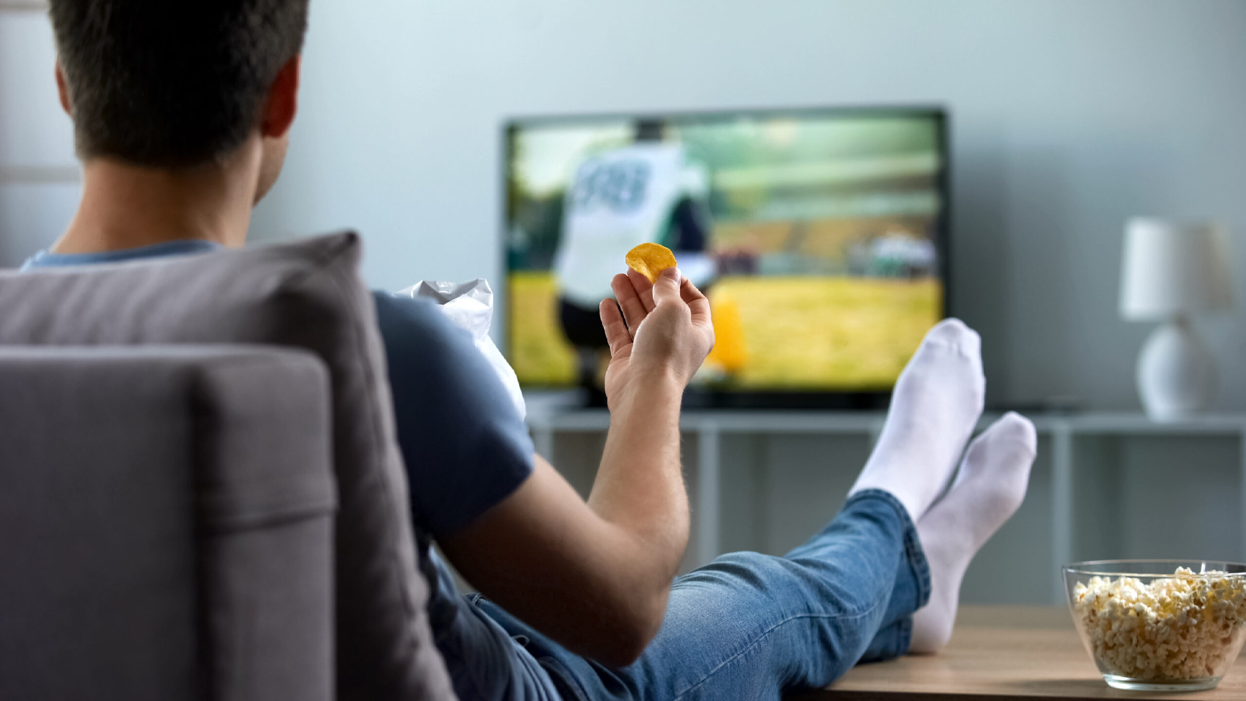 Man watching american football eating snacks on couch at home