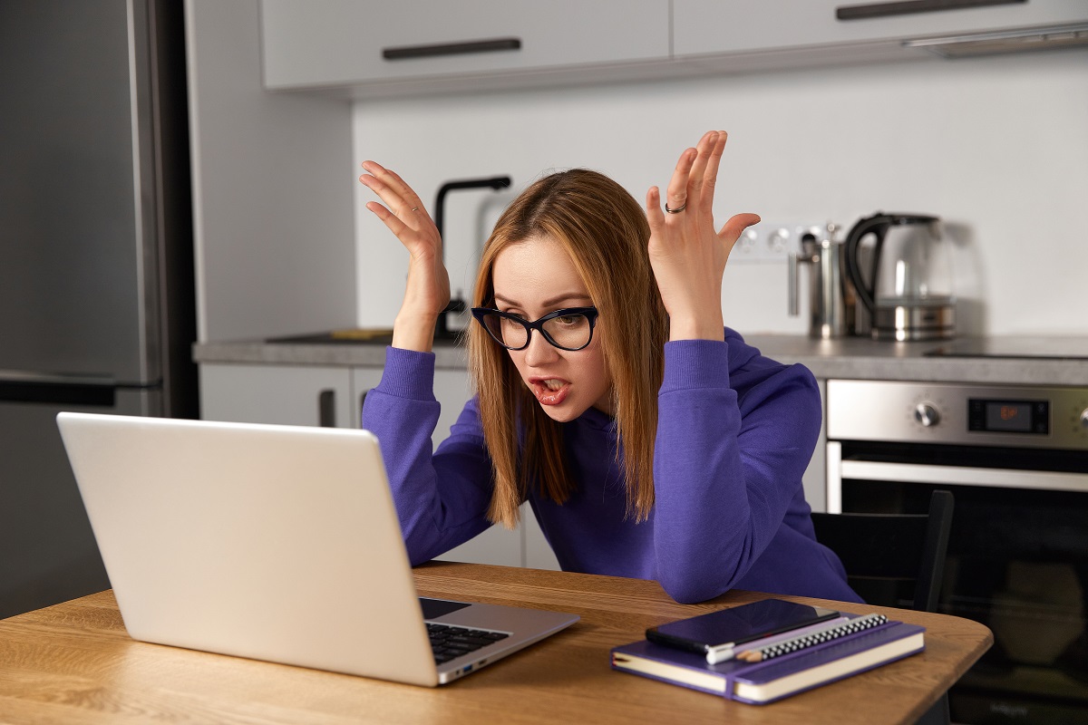 Annoyed young adult woman in glasses using laptop in the kitchen at home