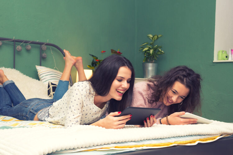 Two smiling young women using smart phone and tablet while lying in bed