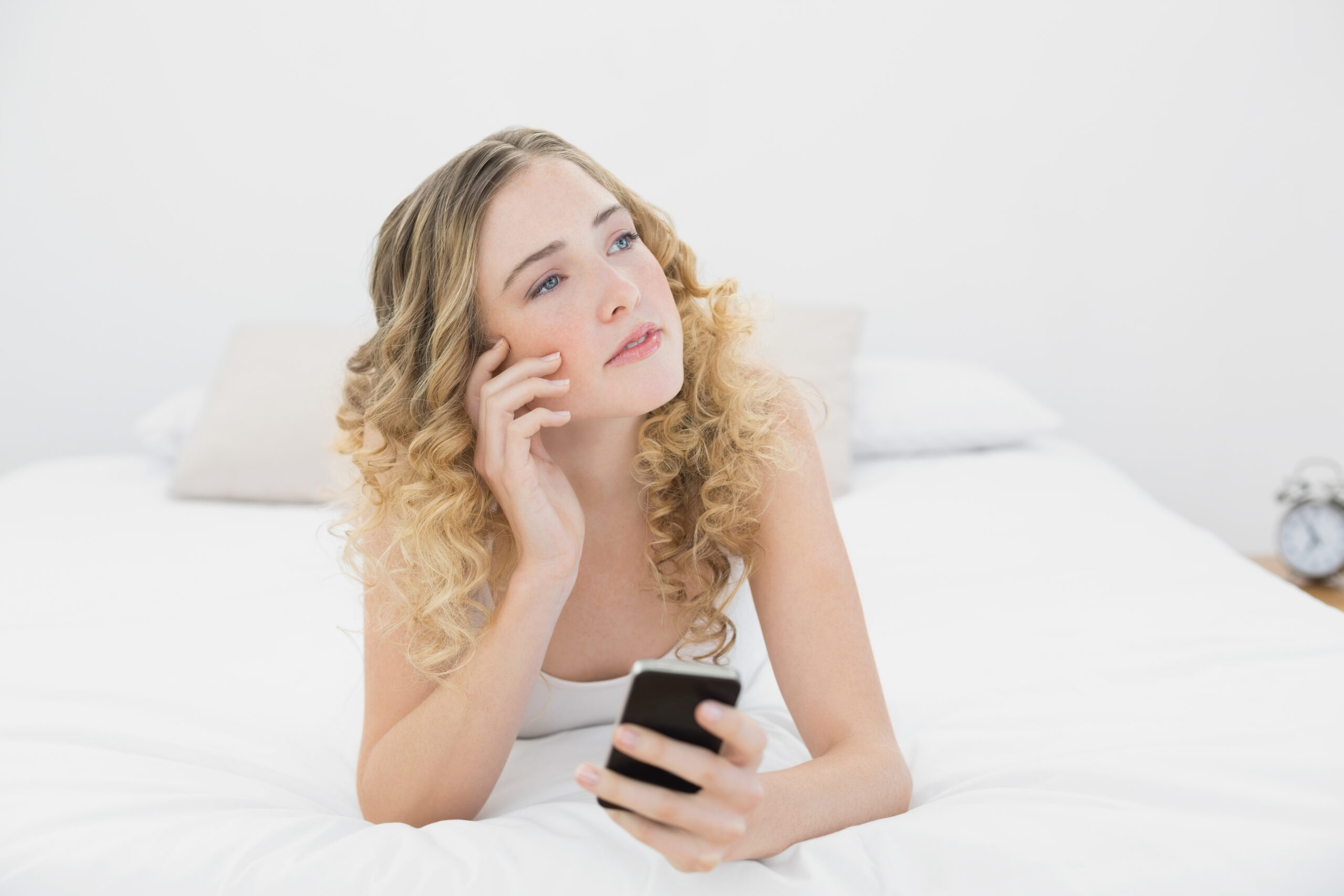 thoughtful blonde lying on bed using smartphone in bright bedroom