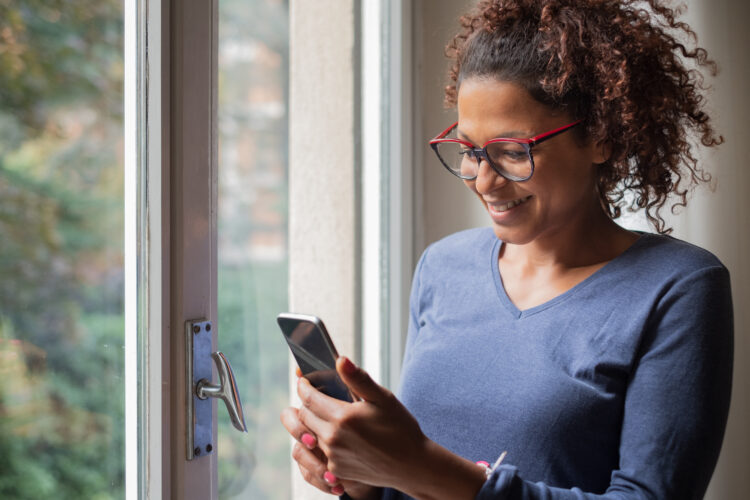 smiling black woman standing beside window while using smartphone