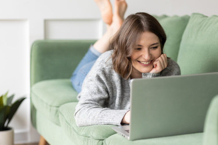 Wanita muda yang cantik tersenyum menggunakan laptop berbaring di sofa