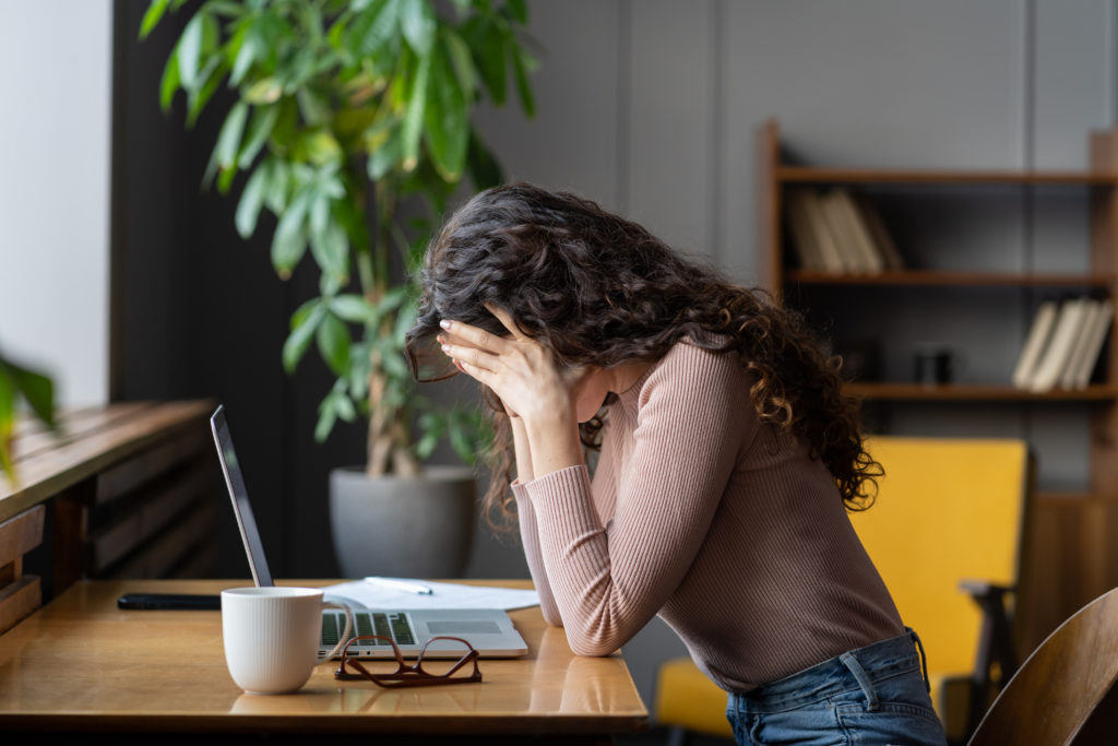  Anxious woman sits at desk with laptop hides face in hands.