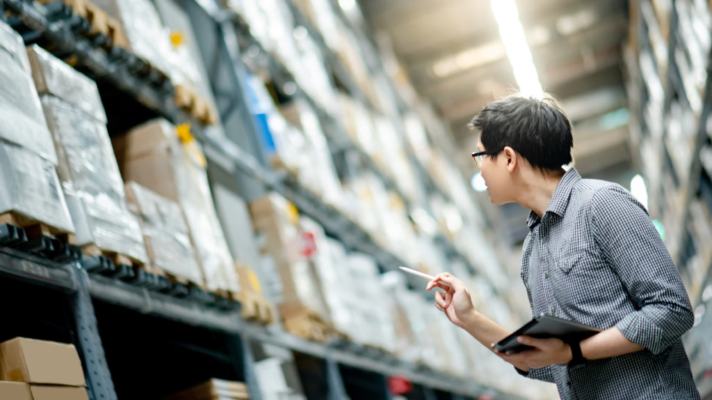 Young Asian man worker doing stocktaking of product in cardboard box on shelves in warehouse by using digital tablet and pen. Physical inventory count concept.