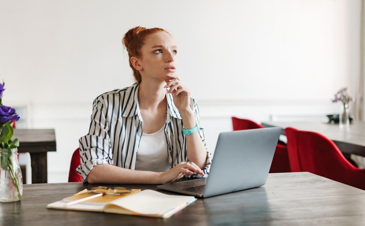 Woman with red ahir in a bun thinking deeply as she works on her laptop