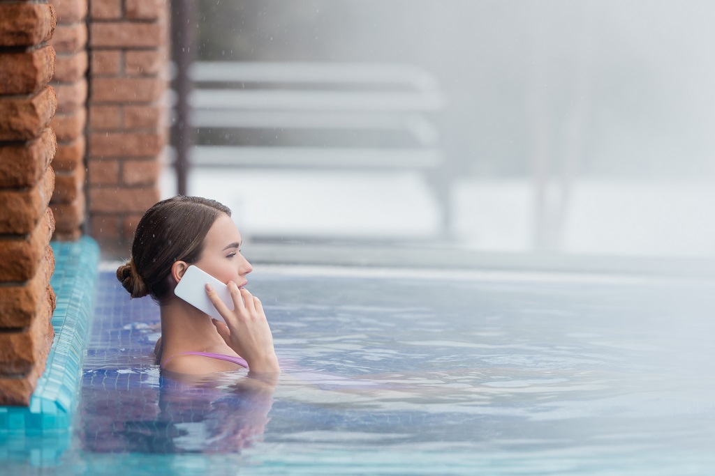 Side view of woman in swimsuit talking on smartphone while bathing in steam bath.