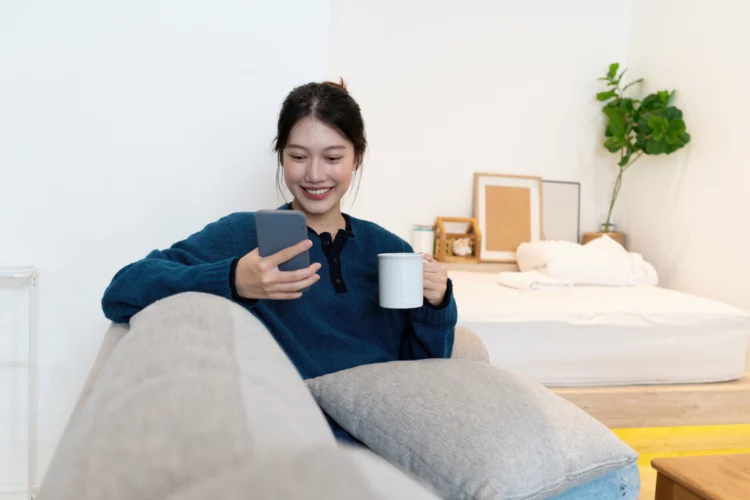 Young Asian woman chatting with her friend and checking social media by smartphone sitting on couch. 