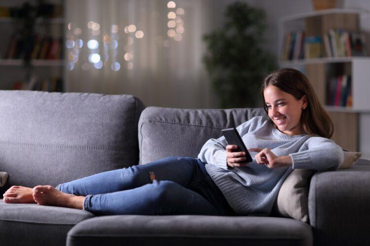 Happy woman using phone lying in the sofa at night at home