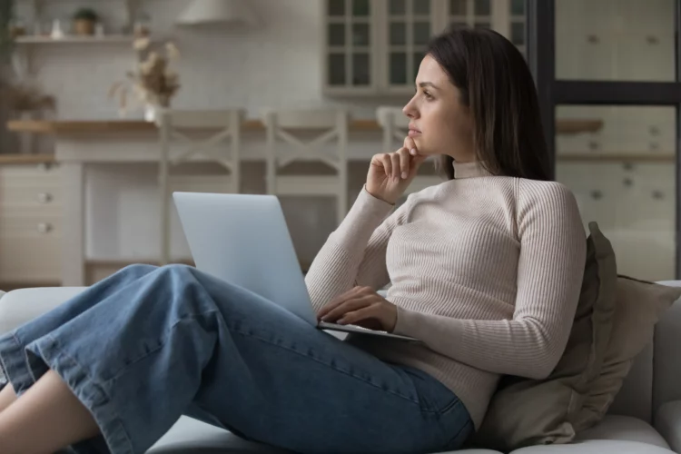 Thoughtful woman sit on couch with laptop staring away