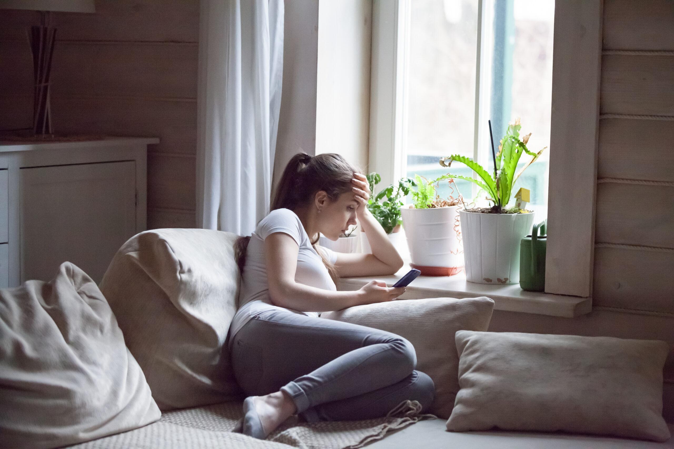 Woman upset after reading bad message on phone at home. Frustrated sad female crying, suffering, not in mood, thinking, depressed, stressed sitting on comfortable sofa near window holding smartphone